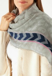 Hellgrauer Schal, der über ein weißes Oberteil drapiert ist, mit einem marineblauen Zickzackmuster und pinken Akzenten, weichem, strukturiertem Finish und fransen gesäumten Kanten.