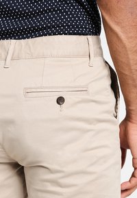 Beżowe chinosy z kieszenią z tylu zapinaną na guzik, przeszyciami oraz dopasowanym krojem. Gładka faktura z prostym, klasycznym stylem.