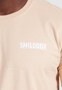Beige katoenen t-shirt met een ronde hals, voorzien van witte reliëftekst "SMILODOX" op het linkerborstgebied. Soepele textuur.