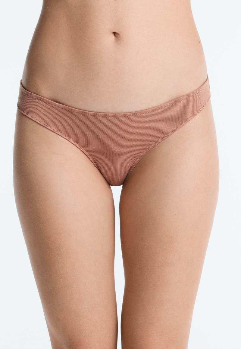 Buste féminin portant un sous-vêtement style bikini côtelé de couleur marron clair sur un fond blanc uni.