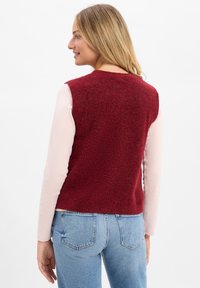 Czerwony dzianinowy kamizelka o teksturowym wykończeniu, noszona na jasnoróżowej bluzce z długim rękawem, w zestawie z niebieskimi dżinsami. Zaprezentowany widok z tyłu.