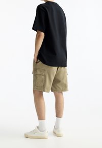 Camiseta de algodón negra, corte holgado, de manga corta, combinada con pantalones cortos de carga beiges con bolsillos laterales. Zapatillas blancas y calcetines completan el look.