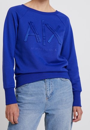 Osoba nosząca jaskrawo niebieską bluzę Armani Exchange z teksturowanymi literami "A|X" i jasnoniebieskie dżinsy.