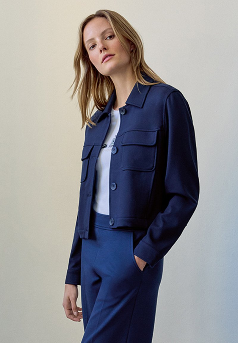 Marineblaues, tailliertes Jackett mit Knopfleiste, zwei Brusttaschen und glatter Textur. Kombiniert mit einem weißen T-Shirt und passenden Hosen.