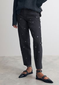 Schwarze Denim-Hosen mit silbernen Ösen, gerader Schnitt, kombiniert mit schwarzen Ballerinas mit Knöchelriemen und dekorativer Hardware.