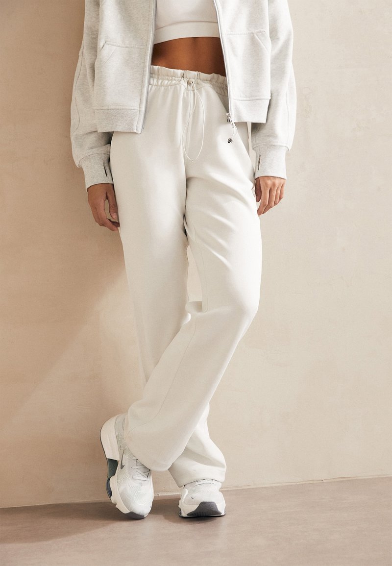 Bílé tepláky s pružn�ým pasem a bočními kapsami, doplněné světle šedou mikinou na zip a bílými sportovními botami.