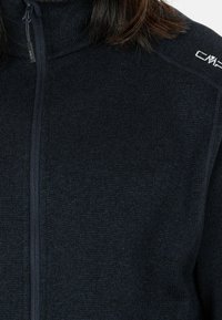 Gros plan d'un pull zippé texturé bleu marine foncé avec logo CMP sur la tirette de la fermeture éclair et sur l'épaule, porté par une personne aux cheveux bruns.