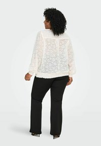 Bijela cvjetna čipkasta bluzica s dugim rukavima i naboranim manžetama, u kombinaciji s crnim hlačama. Gornji dio ima opušteni kroj i strukturiranu obrub.