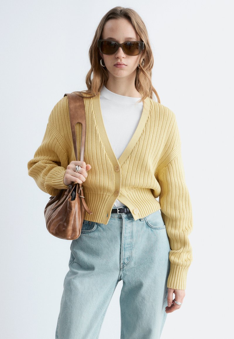 Femme portant des lunettes de soleil marron, un cardigan côtelé jaune, une chemise blanche, un jean bleu clair, et portant un sac à bandoulière en cuir marron.