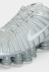 Λευκό και γκρι αθλητικό παπούτσι Nike Shox με πλέγμα στην επάνω πλευρά, ασημί λεπτομέρειες και χαρακτηριστική ελαστική σόλα.