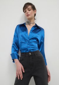 Donna che indossa una blusa arricciata in raso blu lucido, jeans neri a vita alta e una collana choker di grandi fiori d'argento su uno sfondo semplice.