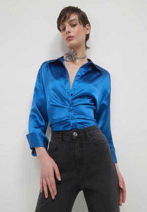 Woman wearing a shiny blue satin ruched blouse, high-waisted black jeans, and a large silver flower choker necklace against a plain wall.
