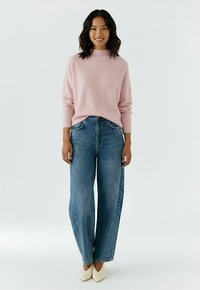 Femme debout portant un pull rose clair, un jean bleu à jambes larges et des chaussures blanches à bout pointu sur un fond clair uni.