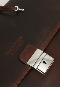 Tummanruskea nahkainen kansio, jossa hopeinen lukko ja nahkahihnaan kiinnitetty pieni avain, painettu teksti "bugatti we are europe".