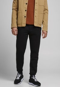 Giacca di tela beige con bottoni neri, indossata sopra un maglione color ruggine. Pantaloni neri slim e sneakers nere con dettagli bianchi e lacci.