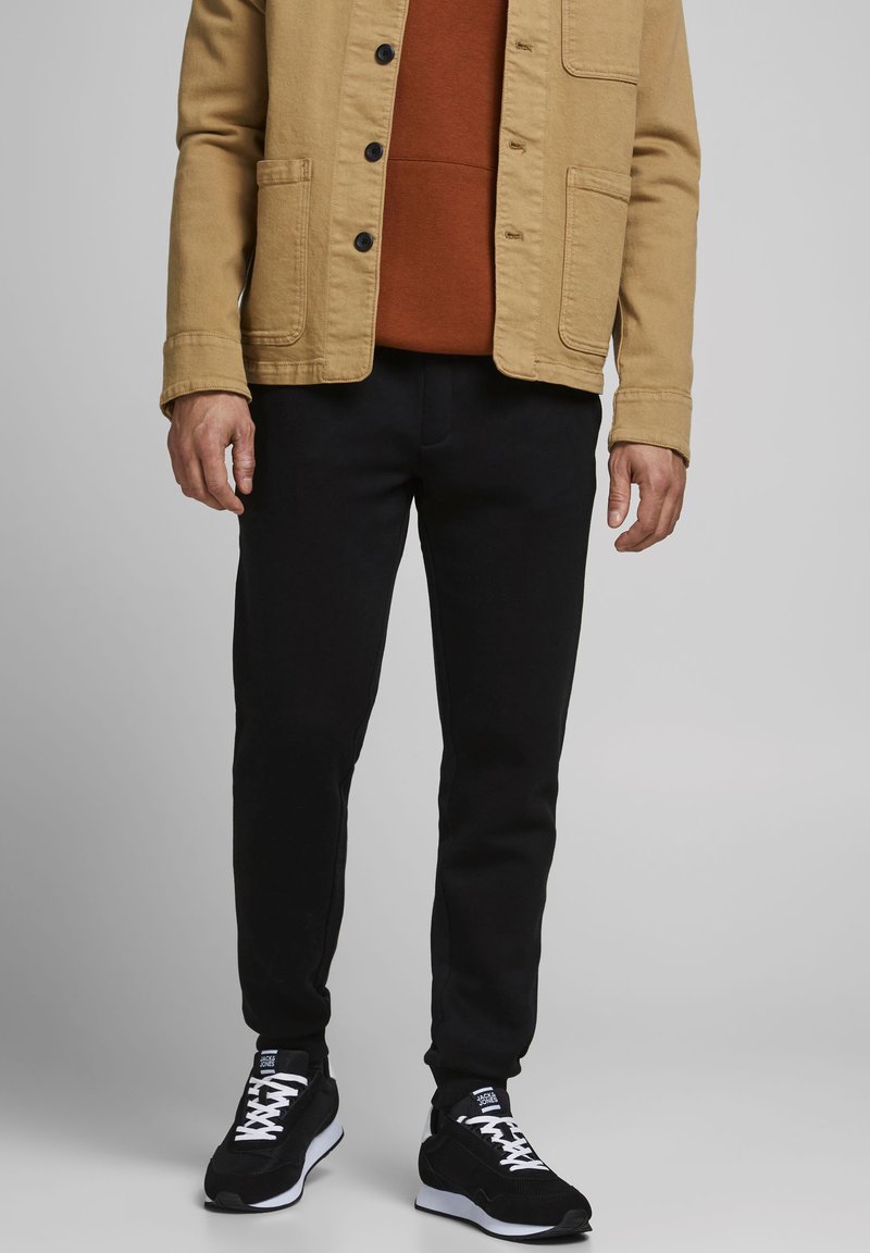Giacca di tela beige con bottoni neri, indossata sopra un maglione color ruggine. Pantaloni neri slim e sneakers nere con dettagli bianchi e lacci.