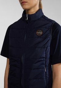 Marinblå puffärmad väst med hög krage, sidogepäck och en rund logotyptryck. Slät textur med vadderade paneler för isolering.