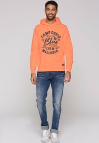 Sudadera naranja neón con estampado gráfico, bolsillo canguro y puños acanalados, combinada con jeans azules enrollados y zapatillas blancas.