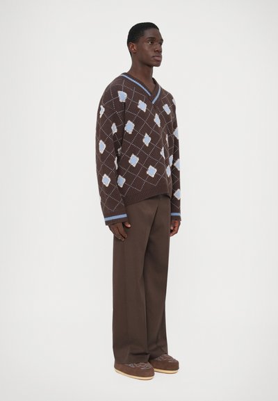 Pulover maro cu un model, având forme de romb în nuanțe de albastru deschis și alb, asociat cu pantaloni maro și pantofi maro cu un design texturat.