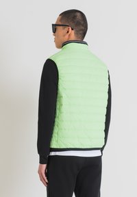 Gilet imbottito verde chiaro con cuciture verticali, colletto e dettagli neri; indossato sopra una maglietta nera a maniche lunghe e pantaloni neri.