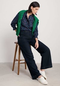 Donkerblauwe blouse met veelkleurige stippen, gerimpelde kraag; groene gebreide trui over de schouders; brede donkere jeans; witte sneakers.