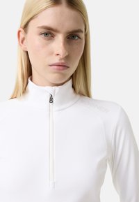 Weißes Sportoberteil mit hohem Kragen und Viertelreißverschluss. Verfügt über dezente Markierung auf der Schulter und glattes, dehnbares Material.