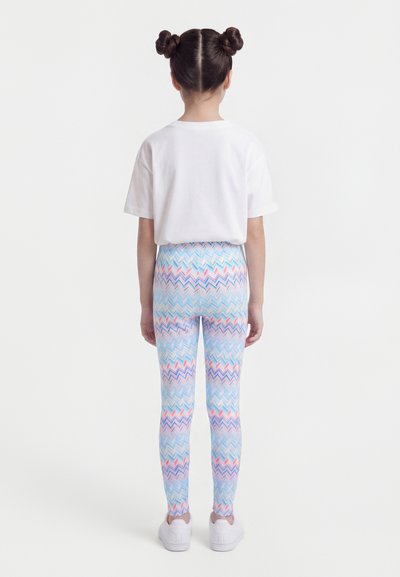 Enfant avec une coiffure à double chignon portant un t-shirt blanc uni, des leggings à motif zigzag coloré et des baskets blanches, debout de dos.