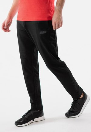 Schwarze Sporthosen mit Seitentaschen und einer glatten Textur, kombiniert mit schwarzen Turnschuhen mit weißen Sohlen und Schnürsenkeln.