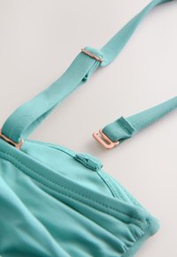 Bretelle de soutien-gorge en tissu turquoise réglable avec curseur et crochet en métal rose doré sur fond clair.