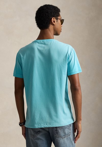 Άντρας που φοράει πετρόλ κοντομάνικο μπλουζάκι, γυαλιά ηλίου και ρολόι, φωτογραφημένος από πίσω σε απλό μπεζ φόντο.