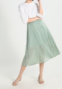 Gonna midi plissettata in verde menta chiaro, con punti metallici. Abbinata a una maglietta bianca e a sandali beige. Tessuto morbido e design fluido.