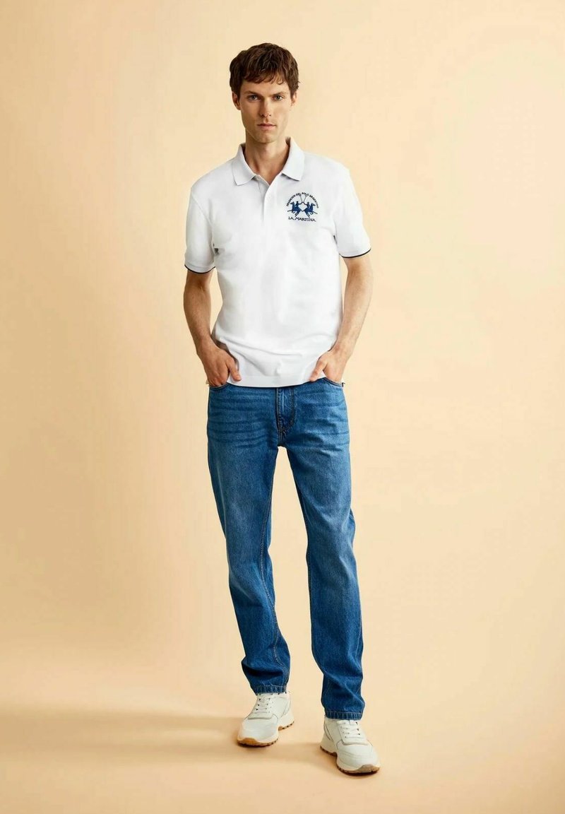 Jonge man staat met handen in de zakken, draagt een wit poloshirt met logo, blauwe spijkerbroek en witte sneakers tegen een effen beige achtergrond.