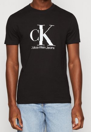 Mężczyzna w czarnej koszulce Calvin Klein Jeans z dużym białym logo "cK" i napisem, w połączeniu z jasnoniebieskimi dżinsami.