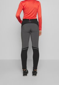 Vysoký pas sportovních legín v šedé a černé barvě s hladkou texturou. Obsahují reflexní detaily a logo na pásku.