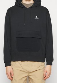 Felpa nera Converse con tasca frontale e cordini, indossata con pantaloni beige, che mostra il torso superiore e le braccia.