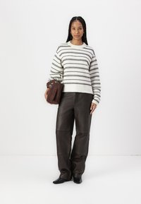 Maglione lavorato a maglia a righe crema e nero abbinato a pantaloni in pelle marroni larghi. Tiene in mano una borsa in suede marrone scuro con una maniglia rettangolare.