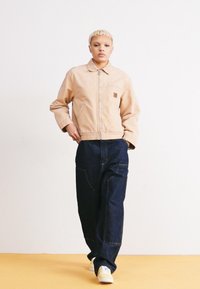 Light beige jacket with a zip closure, side pocket, and logo patch, paired with dark blue wide-leg jeans and white sneakers. Plain background.