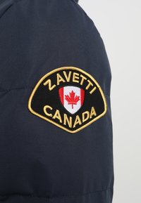 Mørkeblå pufjakke med et trekantet sort og guld patch, der har et rødt ahornblad og teksten "ZAVETTI CANADA" ovenover.