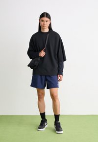 Black oversized sweatshirt, navy shorts, black sneakers, and a small black bag. Background is light with a green floor. Minimalist style.