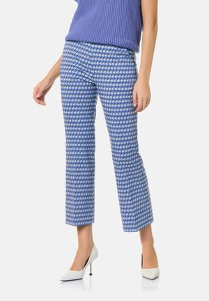 Kobieta ubrana w niebieskie wzorzyste spodnie o długości do połowy łydki, bezrękawnik z niebieskiego dzianego materiału oraz białe buty na wysokim obcasie, stojąca na białym tle.