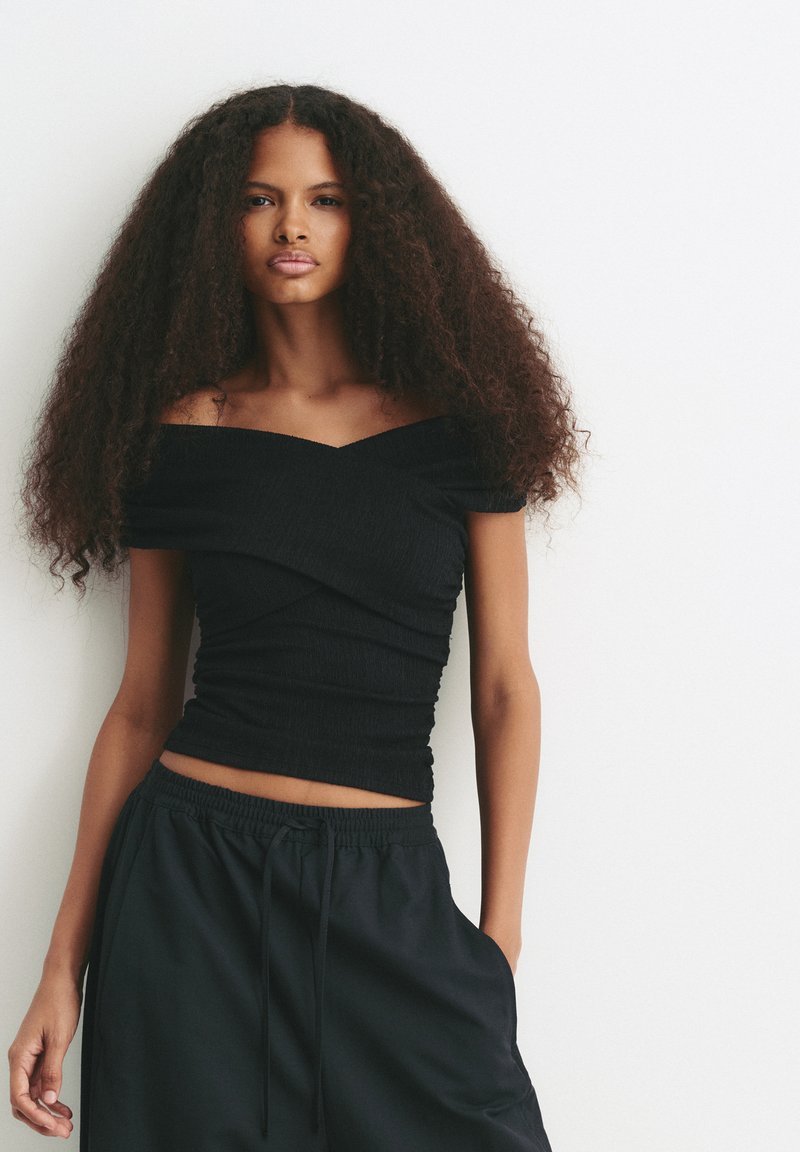 Jonge vrouw met lang krullend haar, gekleed in een zwarte off-shoulder top en zwarte broek, staand tegen een witte achtergrond.