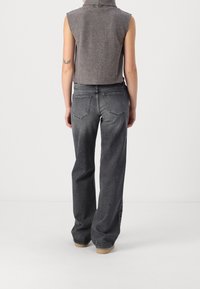 Personne portant un haut gris sans manches et un jean noir délavé à taille haute et jambes larges, debout devant un fond clair uni.