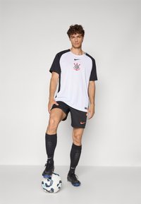Hvit t-skjorte med svarte ermer som har et logo, kombinert med svarte shorts, svarte sokker og svarte fotballsko, stående på en fotball.