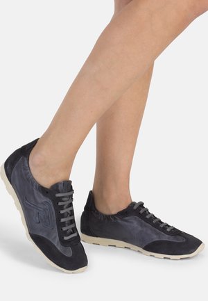 Nogi w czarnych i ciemnoszarych sportowych trampkach na sznurowadła z białymi podeszwami, stojące na jednolitym jasnym tle.