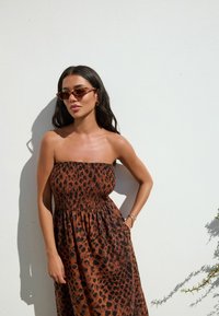 Abito senza spalline con stampa leopardata marrone, dotato di un corpetto arricciato e una gonna fluida. Gli accessori includono occhiali da sole tartarugati e orecchini dorati.