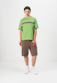 Green T-shirt with gray stripes and logo. Brown cargo shorts with side pockets. White sneakers. Model stands against a plain background.