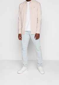 Man in een lichtroze overhemd over een wit T-shirt, lichtblauwe jeans en witte sneakers, staand tegen een effen achtergrond.