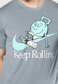 Camiseta gris de manga corta con un gráfico turquesa de un personaje en un rodillo, que presenta el texto "Sigue Rodando". Textura de tela suave.