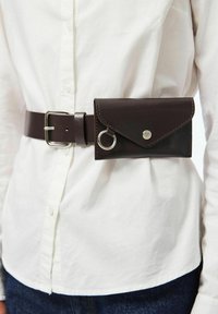 Brūna ādas josta ar pievienotu kabatu, kas aprīkota ar spiedpogu un metāla gredzenu. Gluda tekstūra ar šuvēm gar malām.