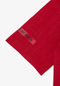 Punainen lyhythihainen paita, jossa on teksturoitu kuvio. Sivussa on erivärinen Lacoste-logopatch.